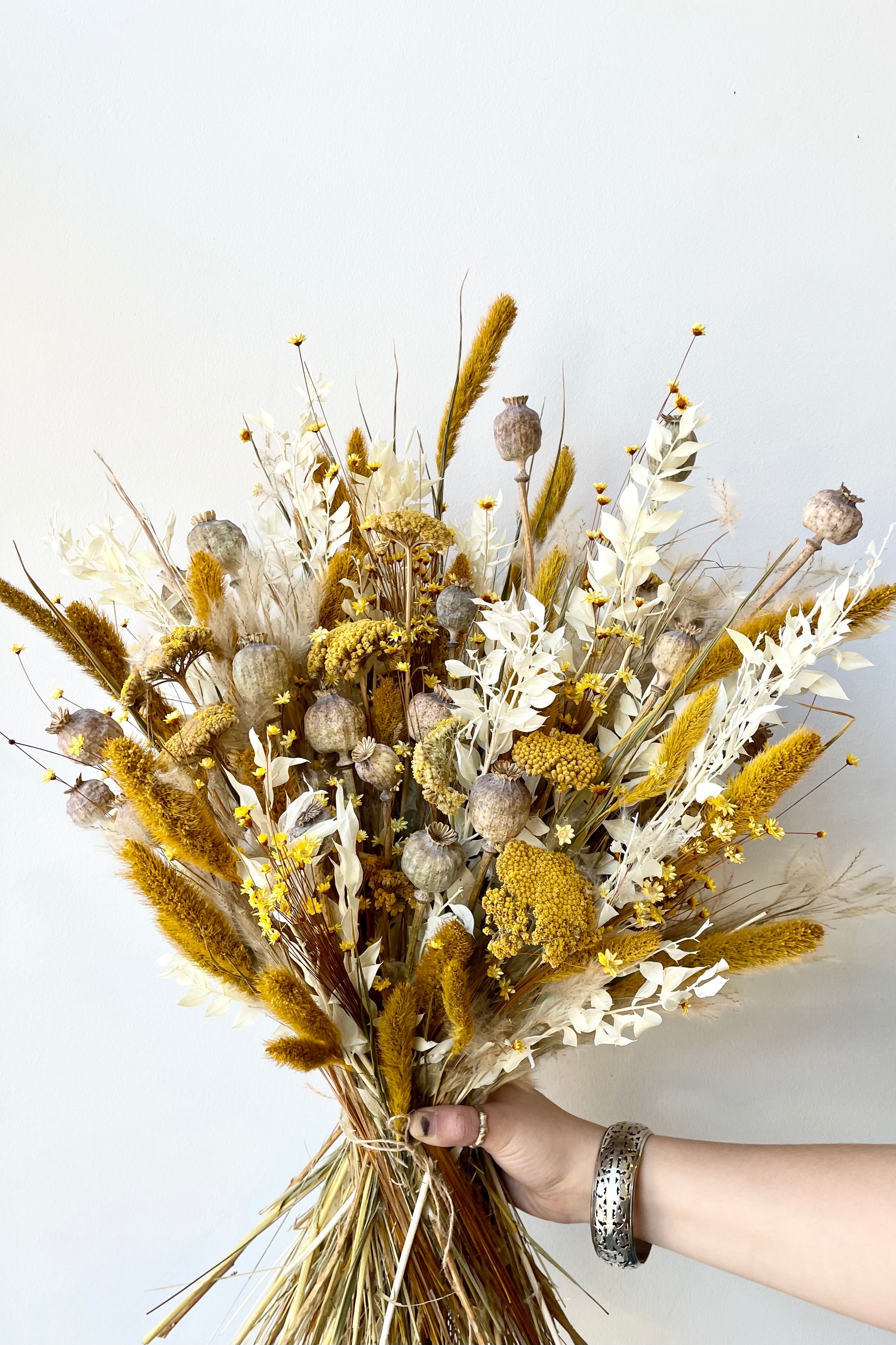 Hand holding a yellow, tan, and white dried floral arrangement with poppies, glixia, yarrow, ruscus, and various grasses. Background is white wall. #size_$200