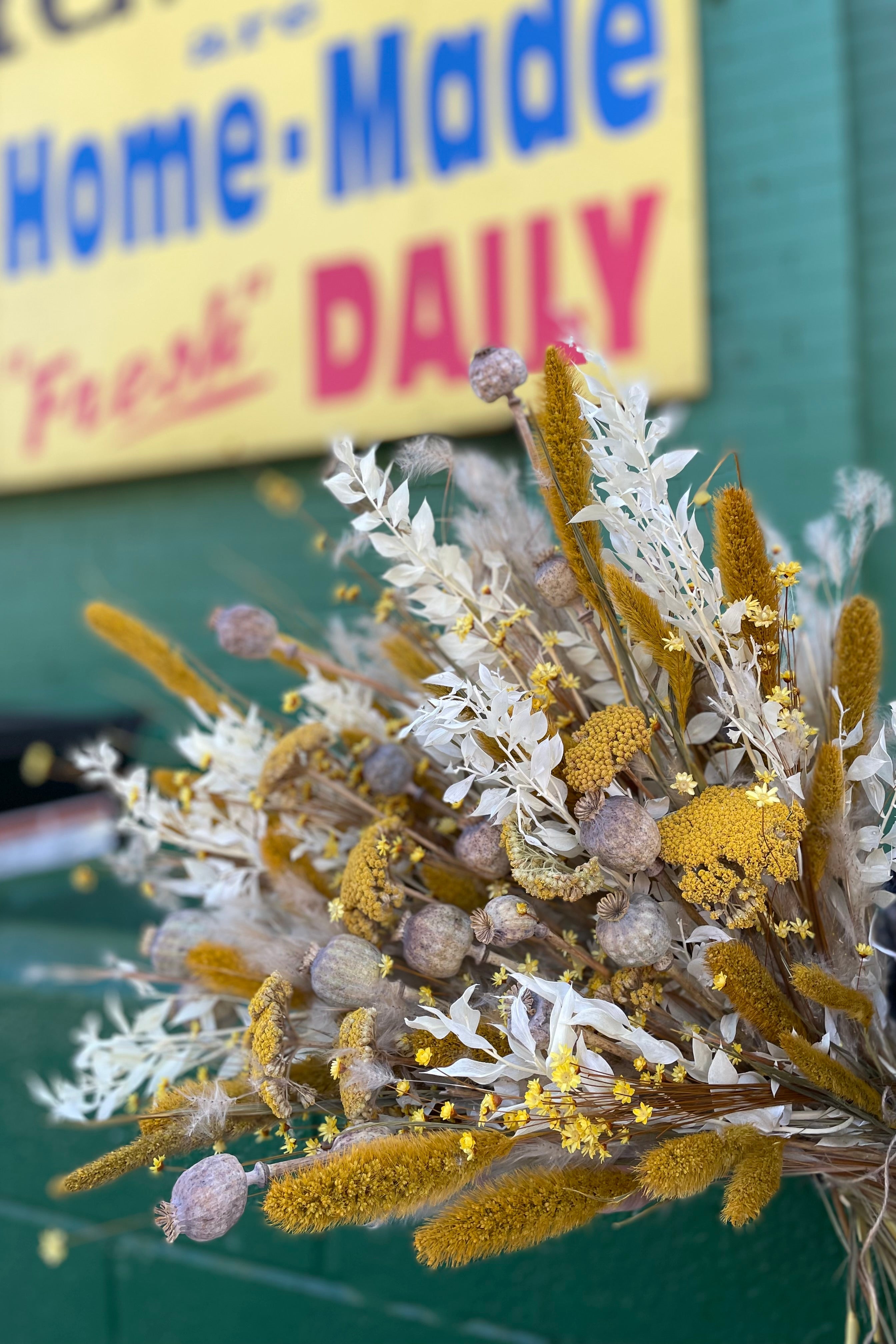 Textures dried floral arrangement with bright colors such as yellow, white and muted tans. All in front of green background and yellow sign with the words "Home Made Fresh Daily".  #size_$200