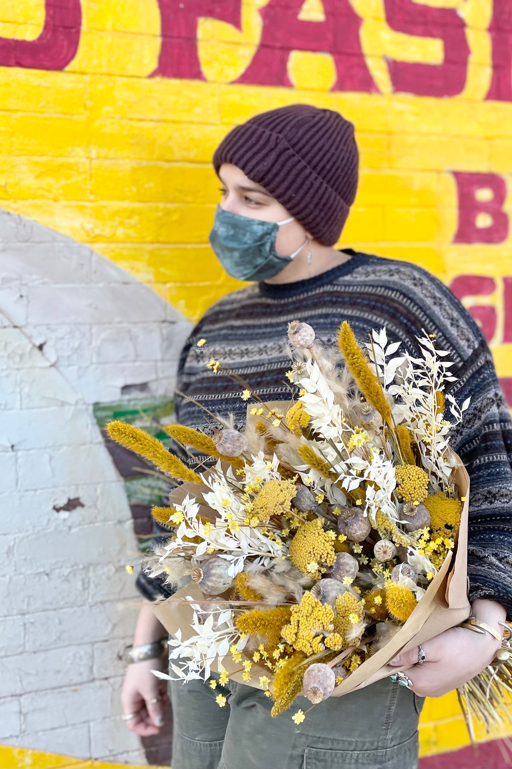 Dried floral arrangement filled with bright colors (yellows, whites and tans) and various interesting textures. Woman holding it against wall. #size_$200