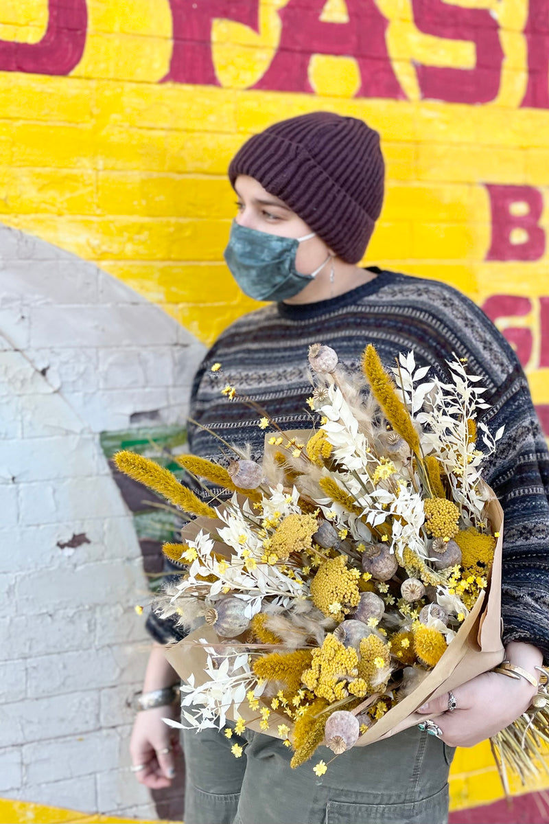 Dried floral arrangement filled with bright colors (yellows, whites and tans) and various interesting textures. Woman holding it against wall. 