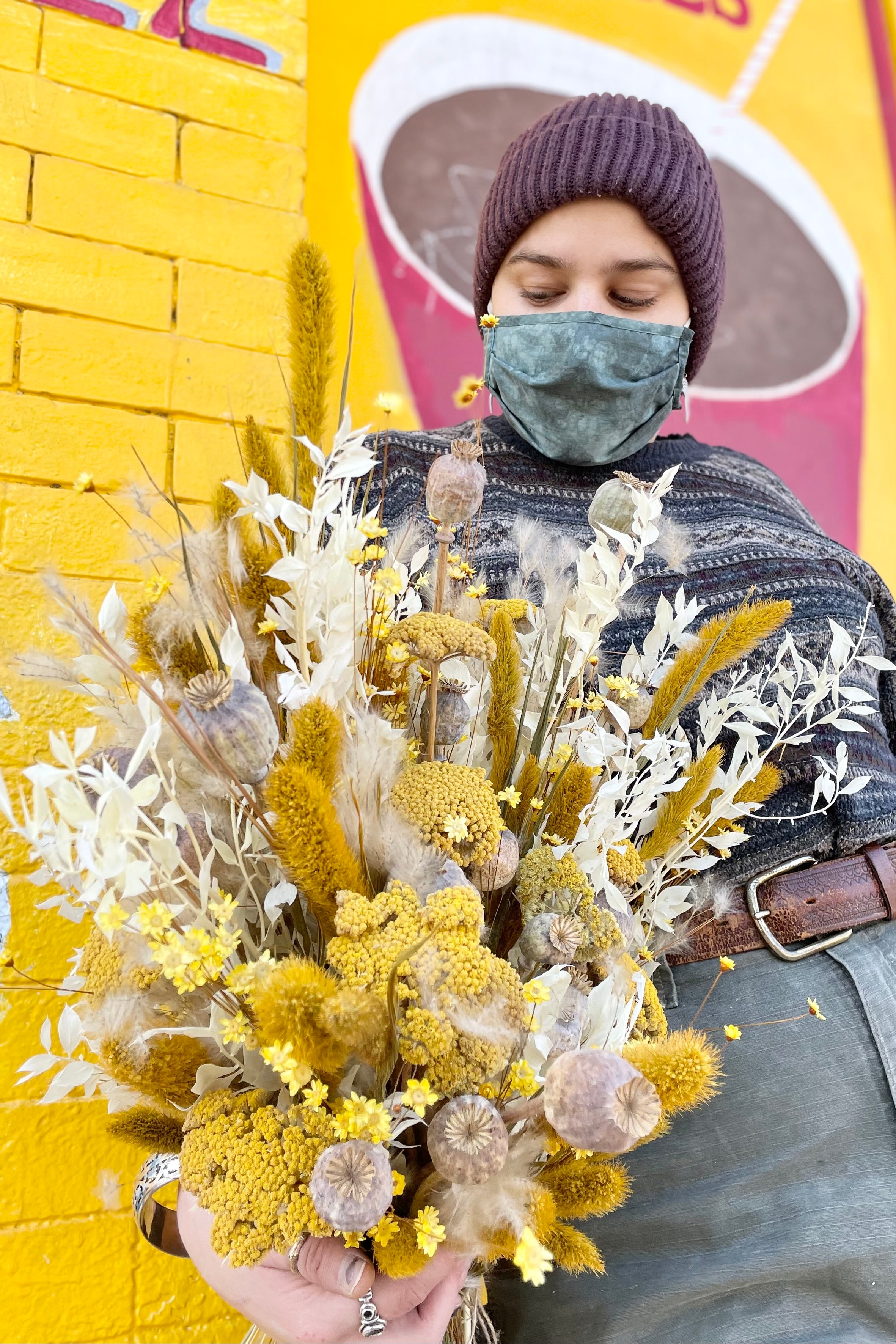 Focal point is a lushes dried floral arrangement of yellow, white, and tan. Comprised of fluffy textures, pods, and grass. Held by person. Background is bright yellow and red. #size_$200