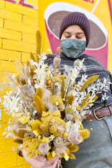 Focal point is a lushes dried floral arrangement of yellow, white, and tan. Comprised of fluffy textures, pods, and grass. Held by person. Background is bright yellow and red.